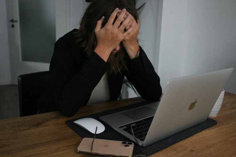 Woman holding her head in stress while sitting at a table with an open laptop, mouse, phone, and glasses in front of her.
