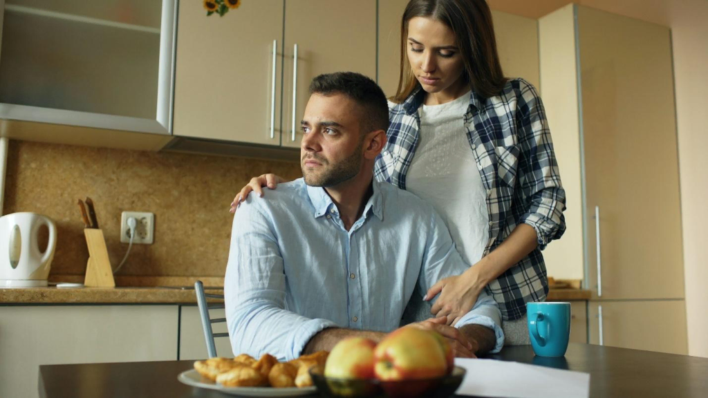 An upset man sitting at the kitchen table while a woman standing by his side comforts him with a hand at his shoulder.