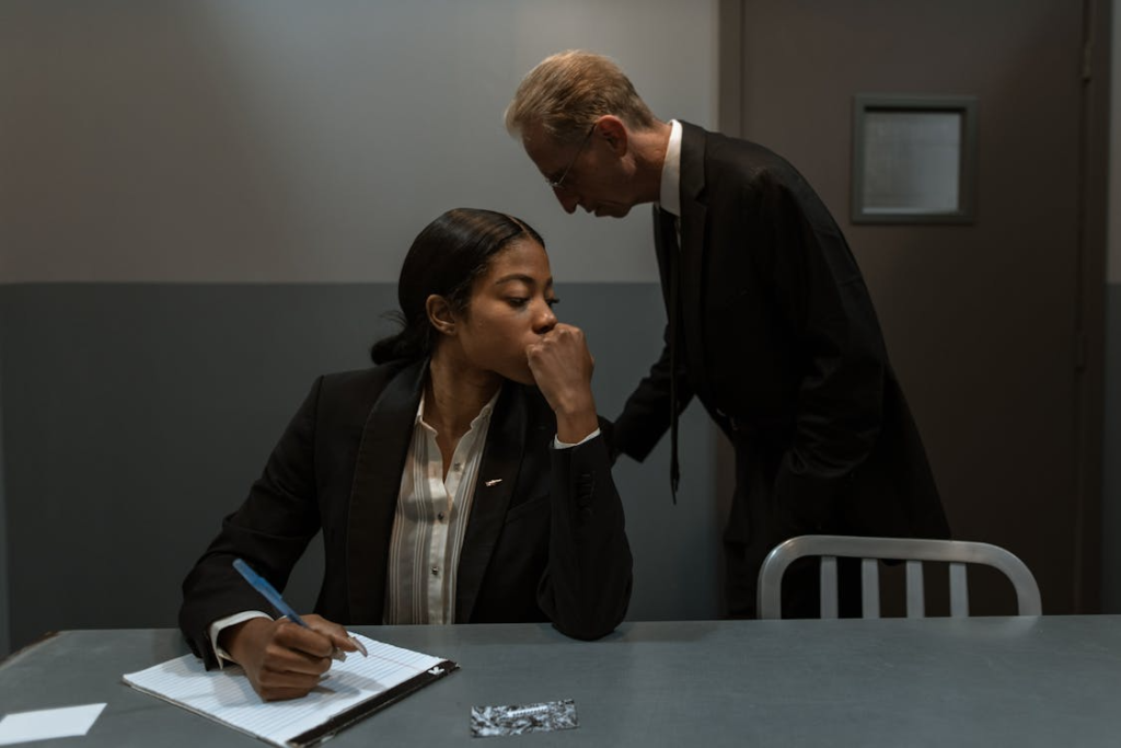 A man in a suit leans over a woman sitting at a table with a notepad, appearing to question her.