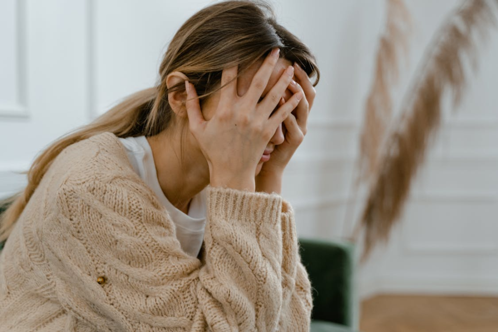 A woman in a beige cardigan sits with her head in her hands, appearing stressed or overwhelmed.